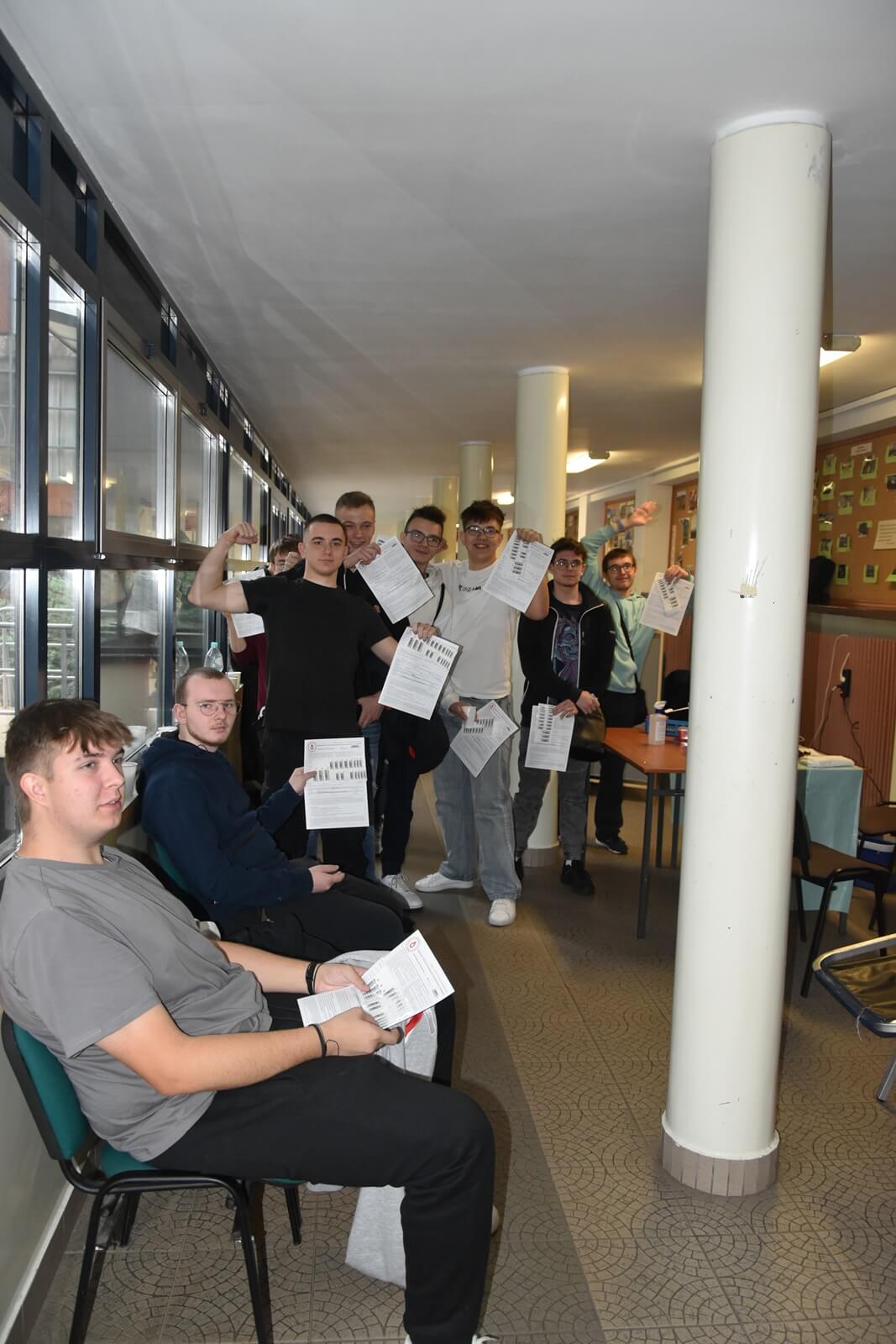 uczniowie z wypełnionymi kartami siedzą i stoją na szkolnym korytarzu czekając na oddanie krwi