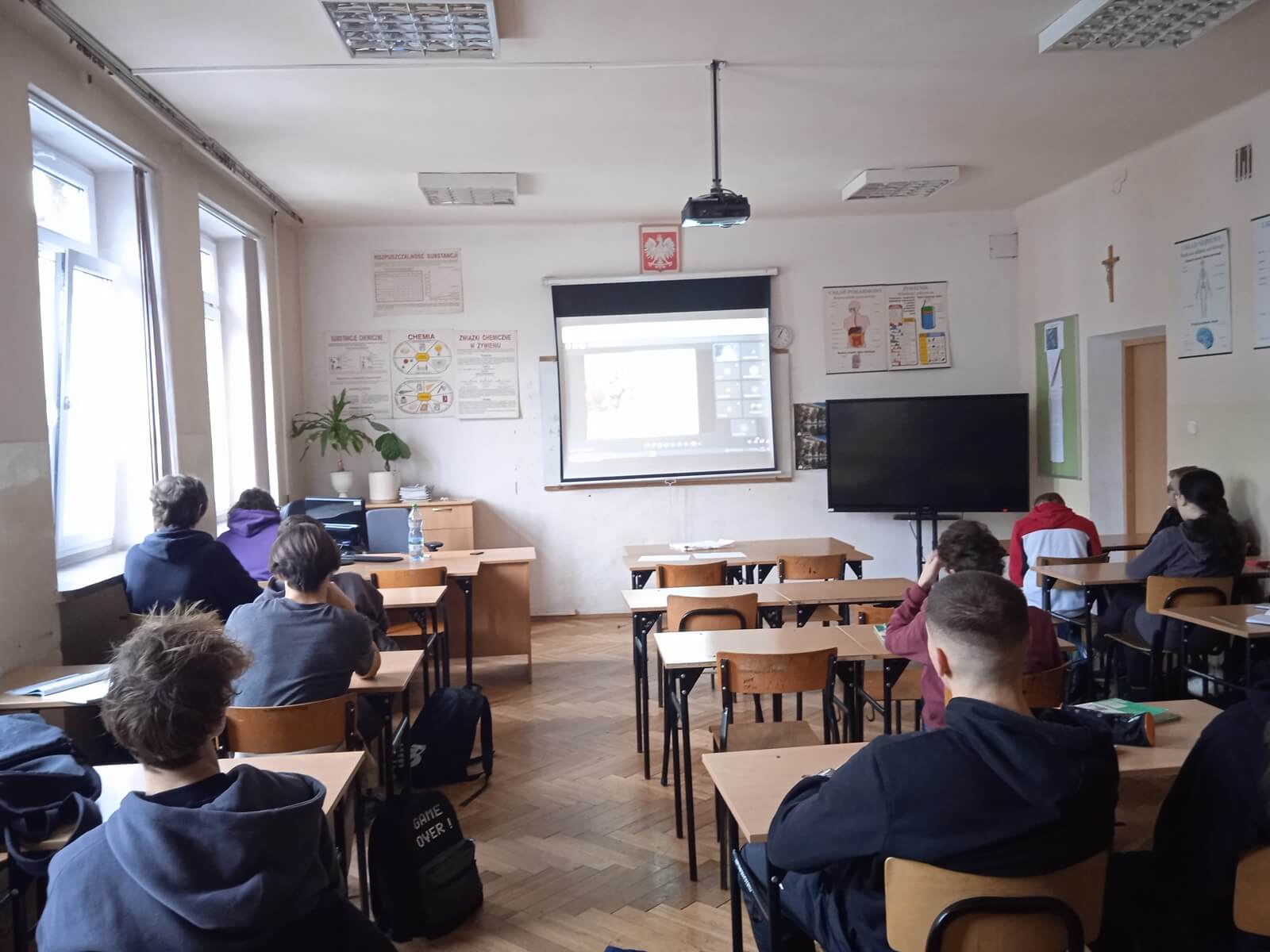 Sala lekcyjna. Uczniowie siedzący w ławkach uczestniczą w zajęciach online