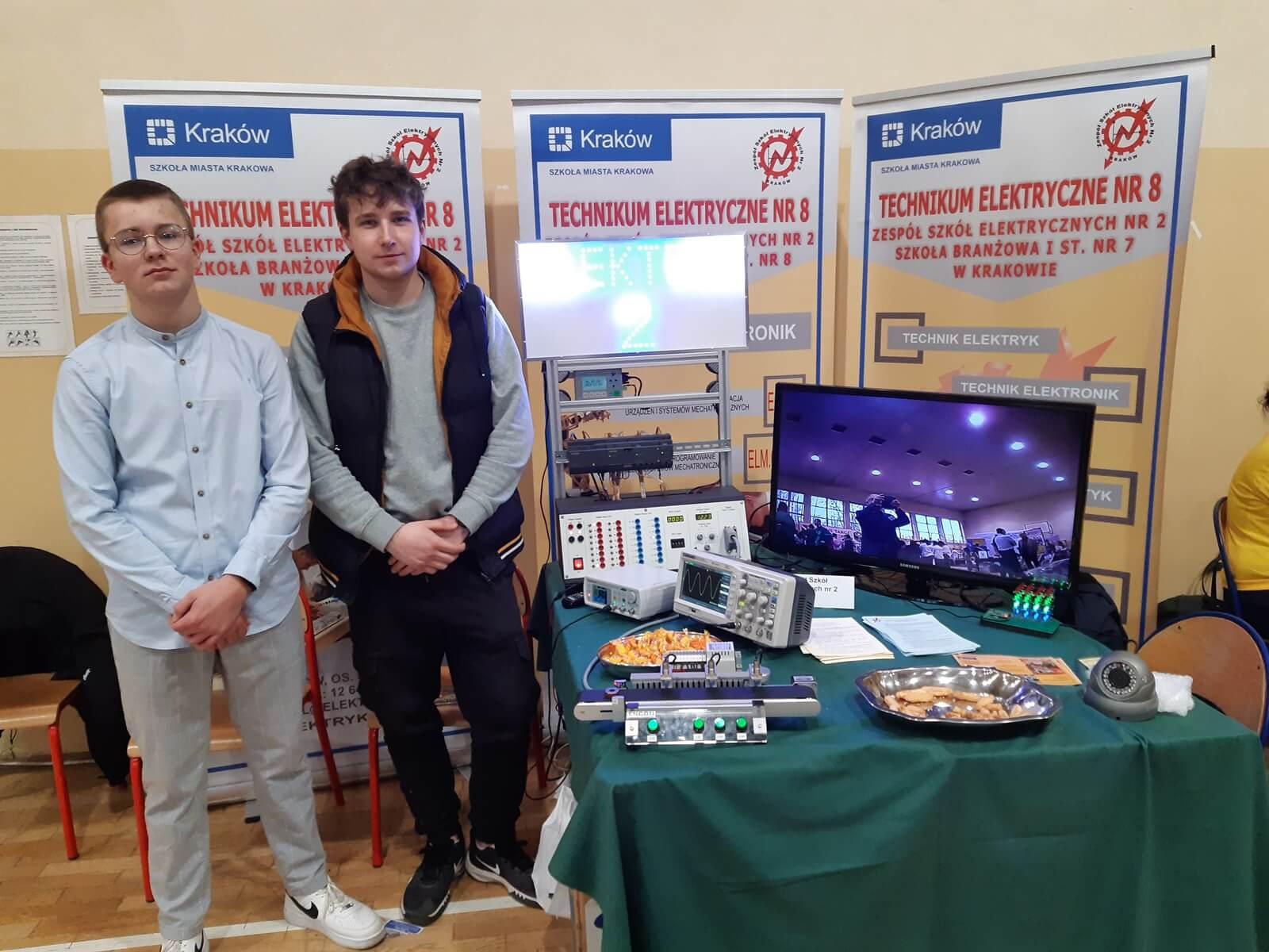Dwaj uczniowie w sali. W tle banery szkoły i stoiki z monitorem, laptopami i przygotowaną prezentacja oferty edukacyjnej szkoły.