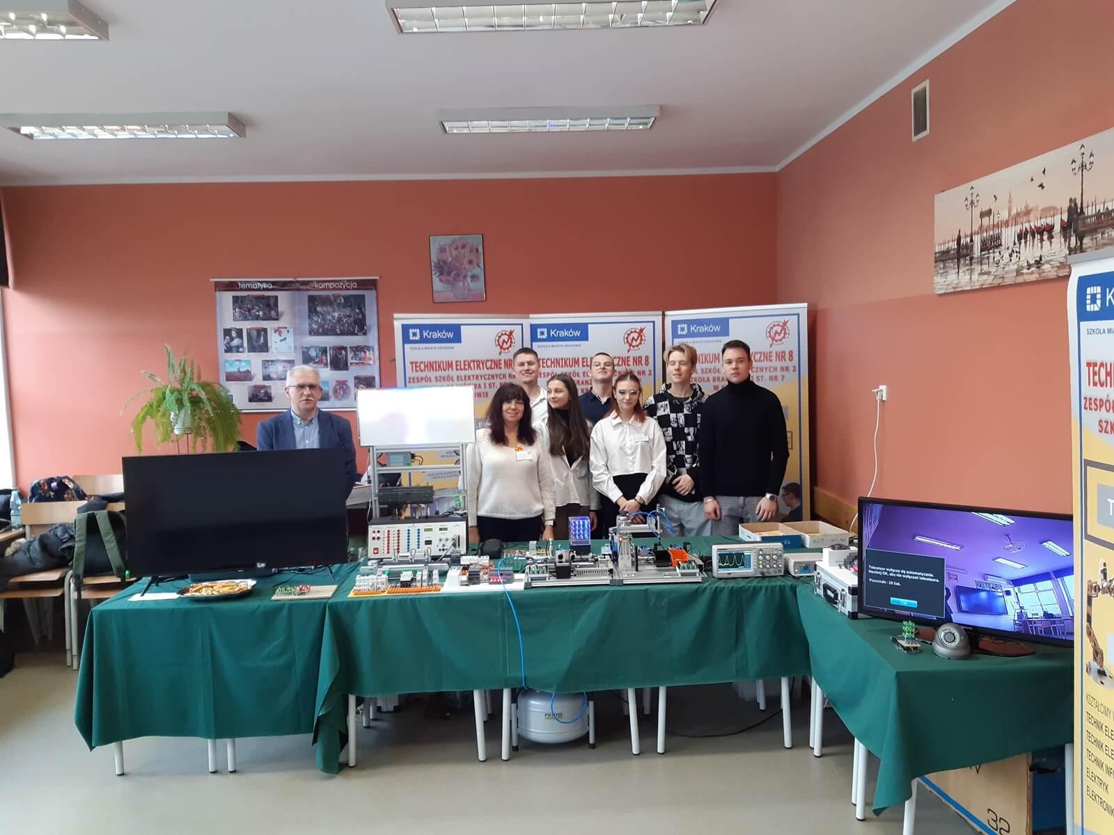 Opiekunowie wraz z uczniowie stoją w sali. za nimi widoczne banery szkoły. Stoisko szkoły w kształcie litery z monitorami, laptopami, ulotkami.