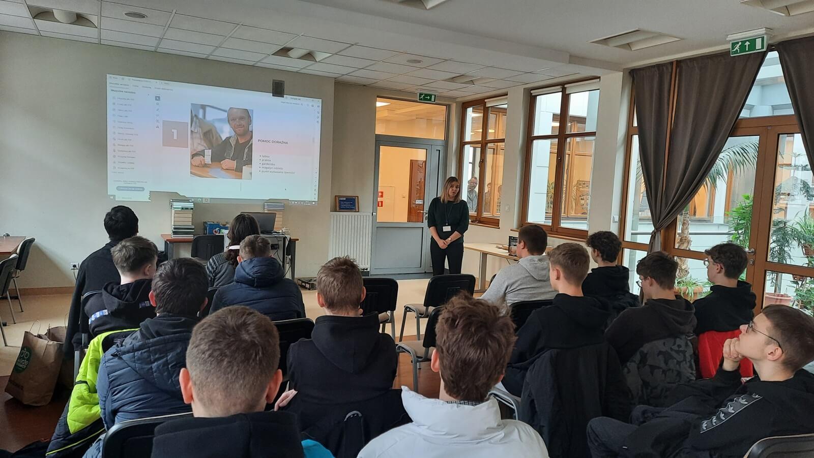 Sala konferencyjna. Przeszklone okna z upiętymi brązowymi zasłonami.. Uczniowie słuchają wykładu i oglądają prezentację.