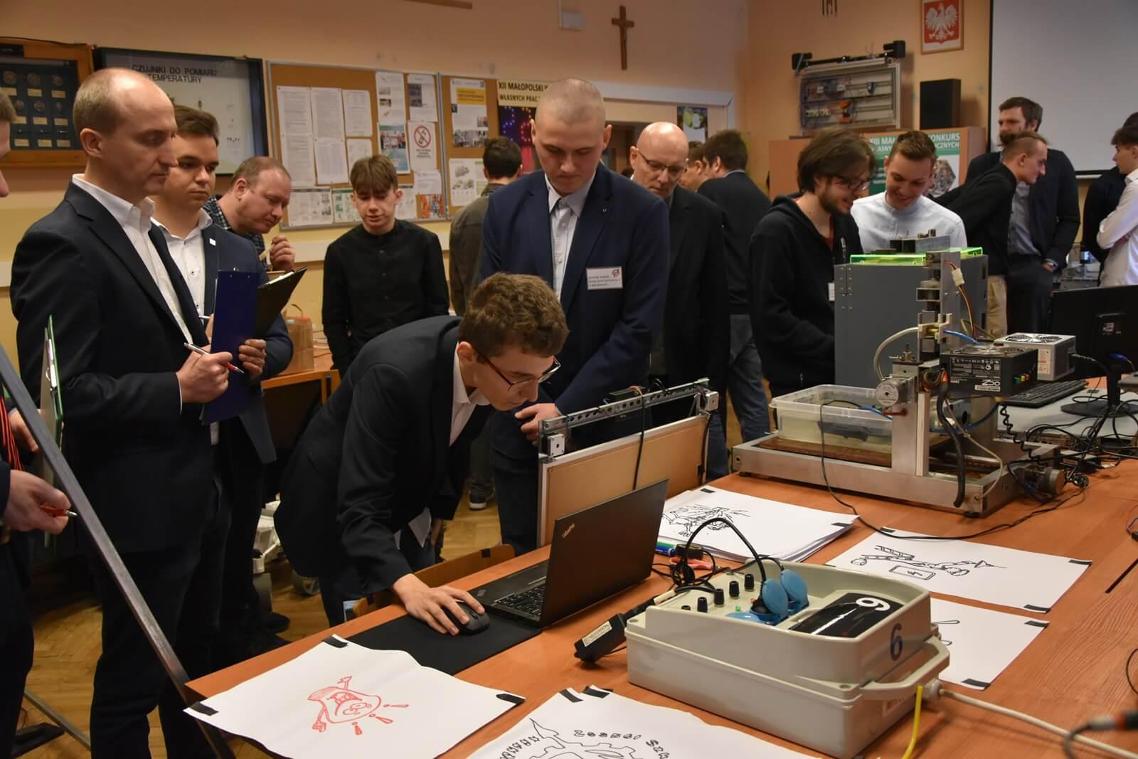 Sala lekcyjna. Na stole stoją prace zgłoszone na konkurs., które oglądają członkowie jury.