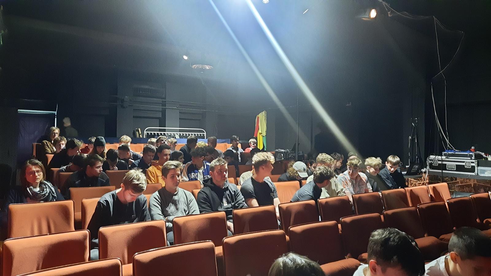 Widownia teatru.Krzesła przykryte tkanina w kolorze pomarańczowym. Widzowie czekają na rozpoczęcie spektaklu.